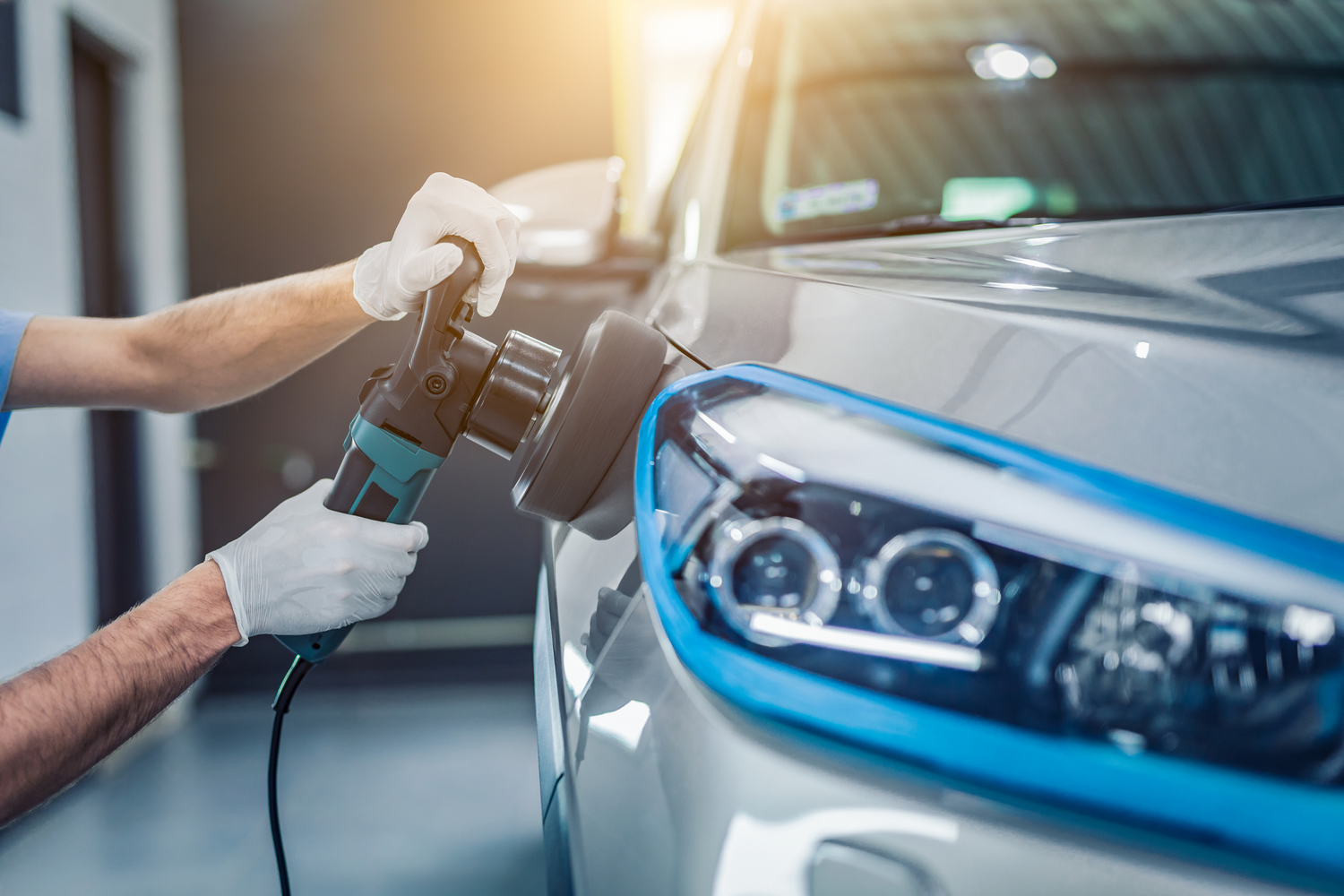 A person wearing gloves uses a power buffer to polish the front headlight area of a silver car, with the vehicle's surface reflecting light.