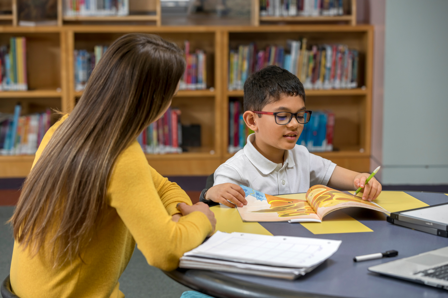 A young boy with glasses reads a picture book at a library table while a woman in a yellow sweater tutors him.